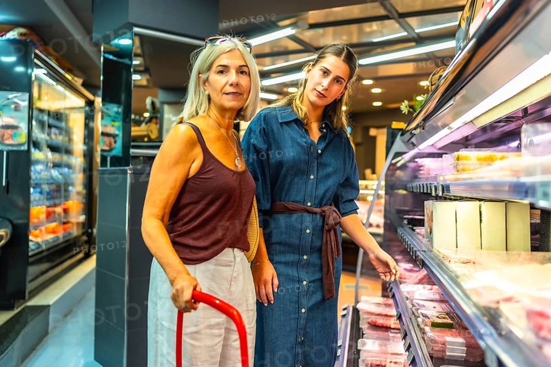 Two women shopping for groceries are choosing meat products from refrigerated section of a supermarket