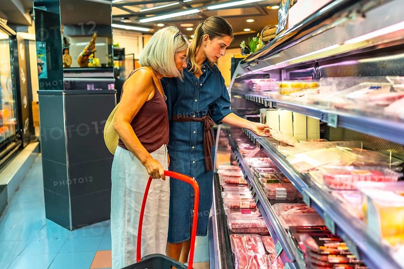 Two women selecting fresh products from a refrigerated section in a grocery store, using a basket for their shopping