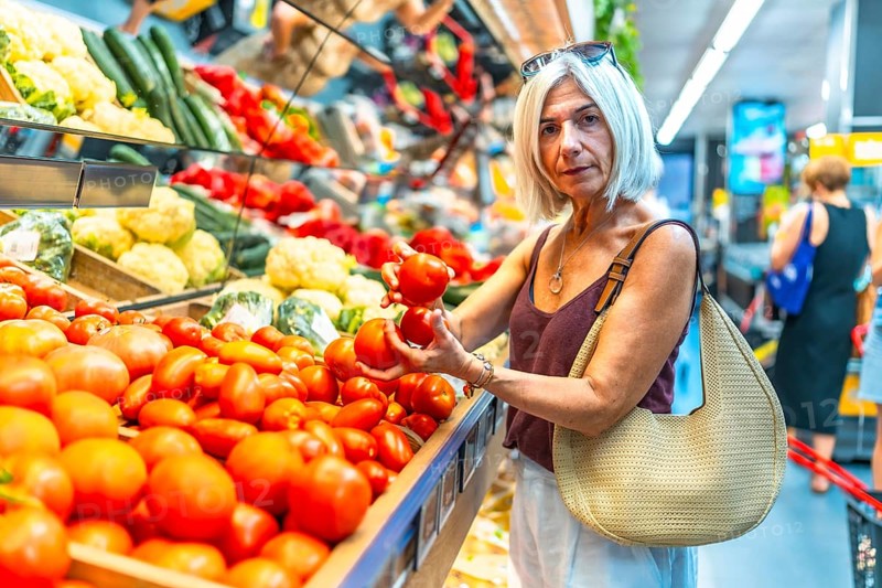 Mature woman choosing ripe tomatoes in a vibrant grocery store produce aisle, surrounded by an array of fresh vegetables and colorful produce