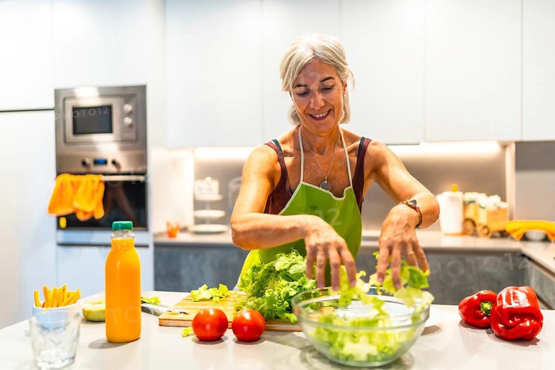 Elderly woman wearing an apron, making a salad with fresh ingredients in a bright kitchen, showcasing healthy lifestyle and cooking at home