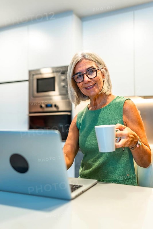 Smiling senior woman enjoying coffee while working on her laptop in a cozy kitchen, embracing a productive and comfortable home office lifestyle