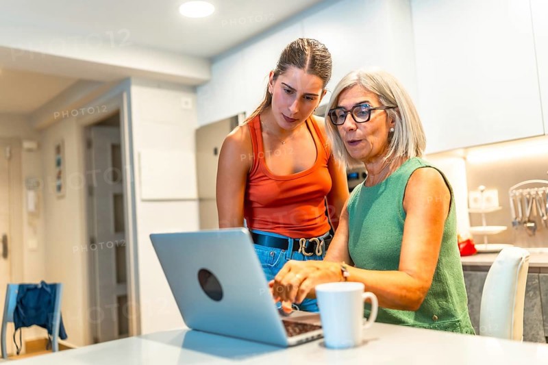 Senior woman navigating a laptop with help from a younger woman in a cozy kitchen, fostering family connection and digital skills