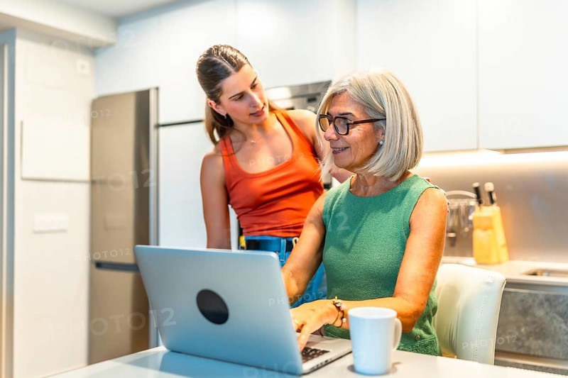 Senior woman is using a laptop in a kitchen while a young woman stands behind her, providing assistance and support