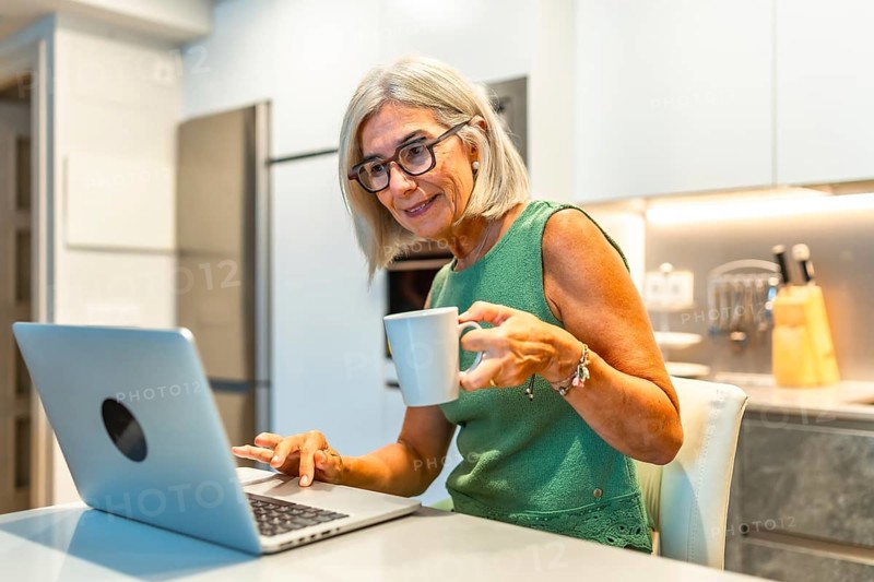 Smiling senior woman using laptop and drinking coffee in modern kitchen, enjoying work from home or retirement