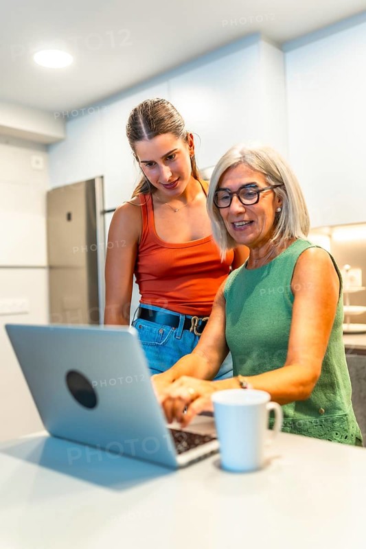 Senior woman using a laptop with guidance from a young woman in a modern kitchen, sharing knowledge and building skills together