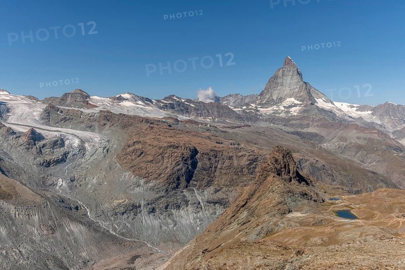 The towering peak of the Matterhorn dominates the clear sky, surrounded by rocky terrain and patches of snow. The barren landscape reveals the natural beauty of the Swiss Alps. Zermatt, Valais