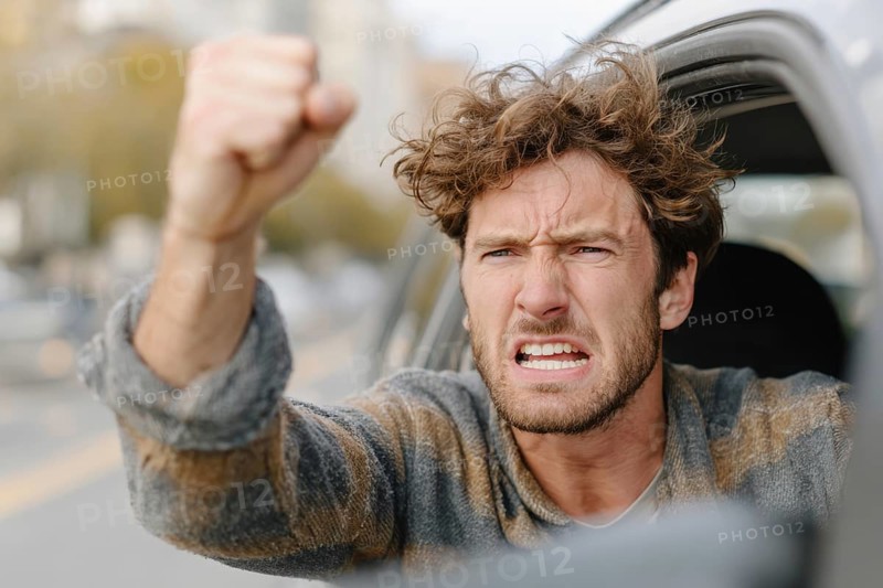 A man in a car shows a clenched fist and looks angrily out of the window on a street, symbolic image for aggressive behaviour in road traffic, AI generated