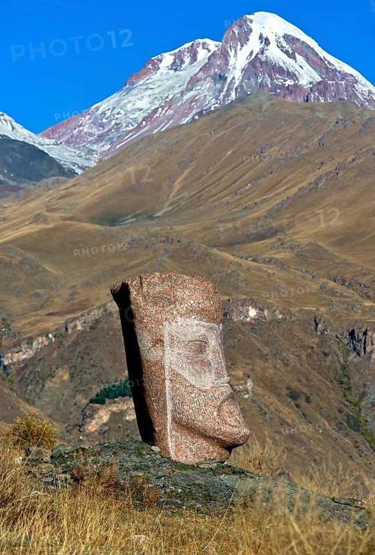 Stone sculpture of a male head, open-air exhibition Stone Heads of Sno by sculptor Merab Piranishvili, the snow-covered peak Kazbek behind, mountain village Sno, Kazbegi municipality in the Mtskheta-Mtianeti region, Georgia