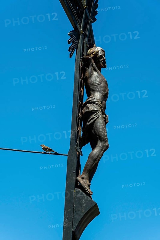 A detailed sculpture depicting Jesus on a cross rises against a bright blue sky. A small bird can be seen perched on a wire, adding life to the serene scene of religion and nature, Puy de Dome, Auvergne Rhone Alpes, France
