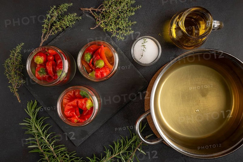 Glasses filled with chilli peppers next to a saucepan with vinegar, oil and herbs on a dark background