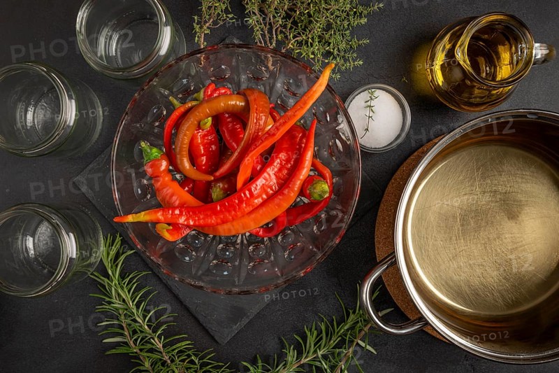 Hot peppers with empty jars, herbs and cooking pot on a dark table