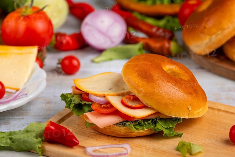 A bagel topped with cheese, sausage, tomatoes, onion and lettuce leaves on a wooden board