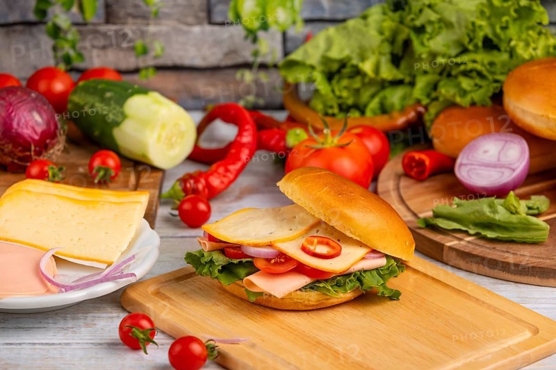 A bagel topped with cheese, sausage, salad and vegetables, surrounded by fresh ingredients