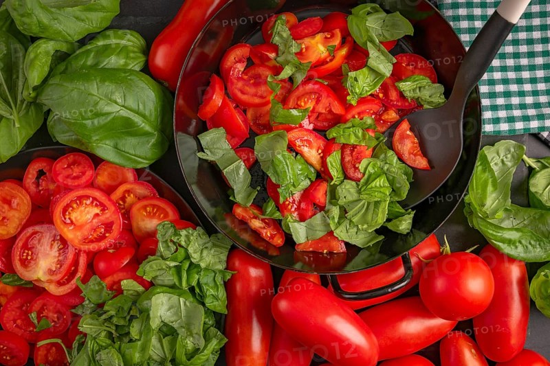 A pan filled with sliced tomatoes and basil leaves, surrounded by fresh vegetables