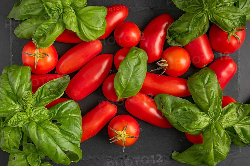 Bright red tomatoes next to fresh basil on a dark slate plate