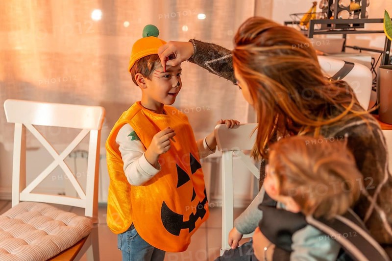 Mother applying halloween makeup on her son wearing a pumpkin costume, while holding her baby