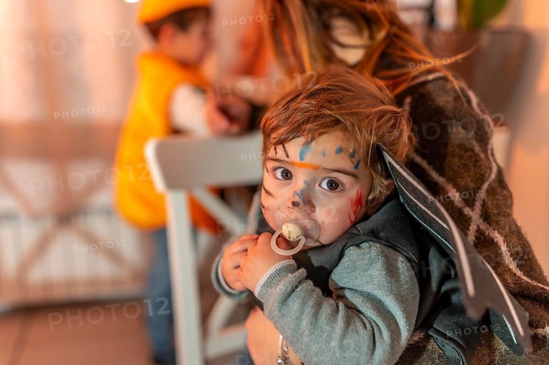 Toddler wearing a halloween costume with painted face and pacifier, held by mother, with another child in the background