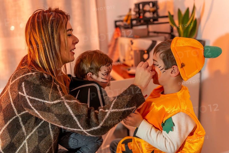 Mom putting halloween makeup on her two sons, dressed up as a bat and a pumpkin, in their home