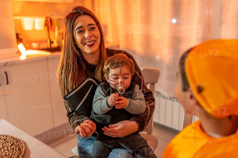 Mother holding her baby dressed up as a bat while smiling at her other child dressed in orange costume, celebrating halloween