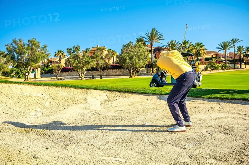 Golfer concentrating on hitting ball out of sand trap on a beautiful sunny day, with golf cart in background