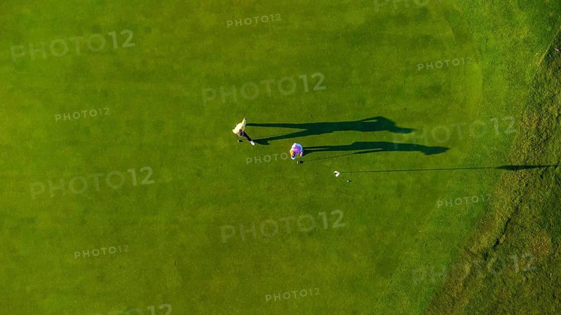 Two golfers are putting on a vibrant green golf course, seen from an aerial perspective, creating long shadows in the afternoon sun