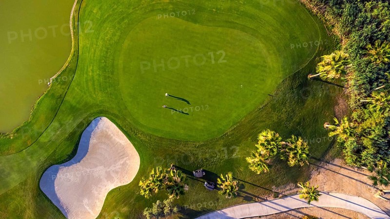 Aerial view of a vibrant green golf course with players enjoying a round, featuring a sand trap, palm trees, and a golf cart