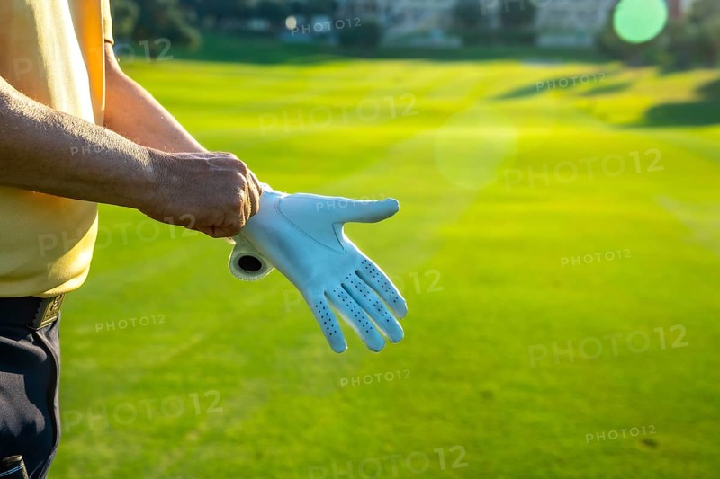 Golfer putting on a white golf glove, getting ready for an exciting game on a lush, vibrant green golf course under the summer sun