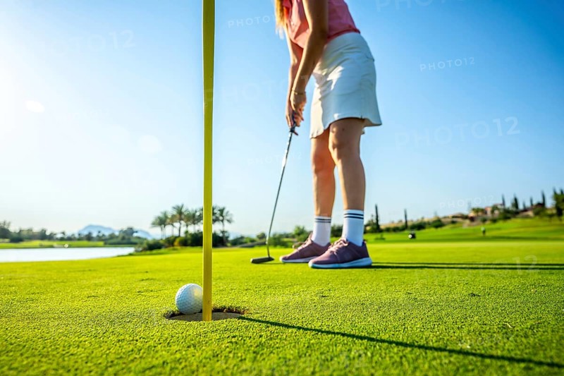 Female golfer putting a ball into the hole on a sunny day, enjoying leisure time on a stunning golf course surrounded by nature