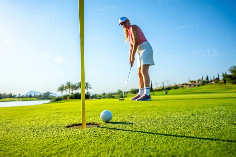 Woman golfer putting a ball on the green, aiming for the hole under a bright sun at a picturesque golf course on a summer day