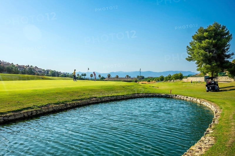 Two golfers are playing on a sunny golf course with a water hazard and golf cart in the background