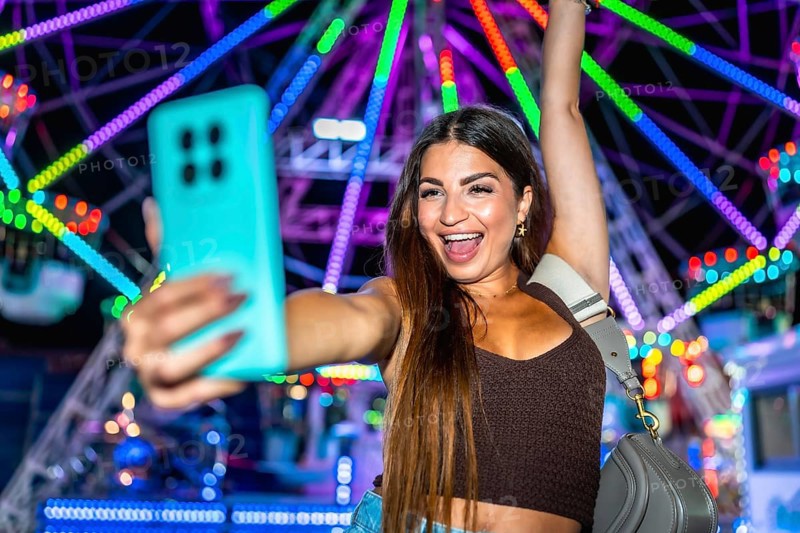 Young woman taking a selfie with smartphone in front of a colorful ferris wheel at an amusement park, enjoying the vibrant atmosphere