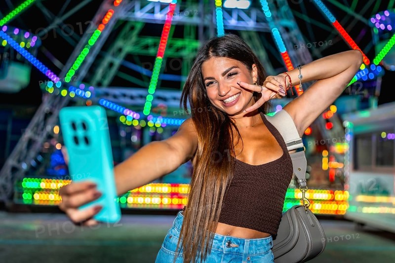 Happy young woman taking a selfie and making peace sign at amusement park with colorful ferris wheel in background at night