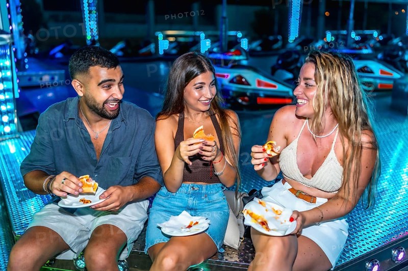 Three friends are enjoying sandwiches while sitting on a bumper cars platform, illuminated by colorful lights at night