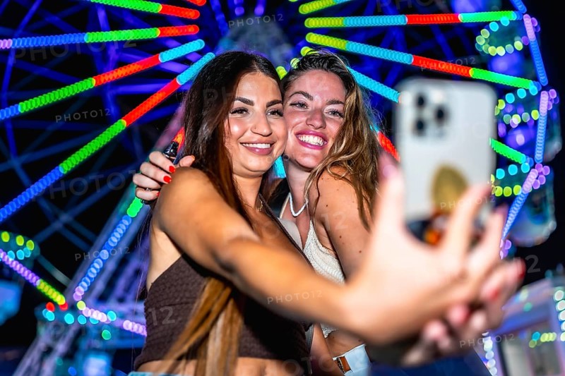 Two friends are taking a selfie in front of a brightly lit ferris wheel at an amusement park at night