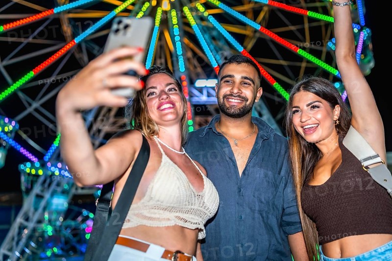 Three happy friends taking a selfie with smartphone in front of a colorful ferris wheel at an amusement park at night