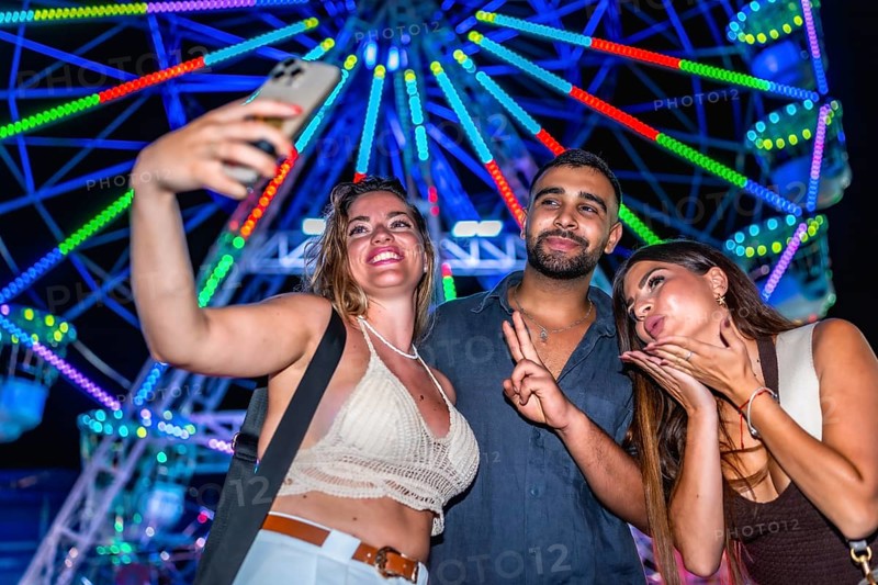 Three friends taking a selfie with a smartphone in front of a brightly lit ferris wheel at an amusement park at night