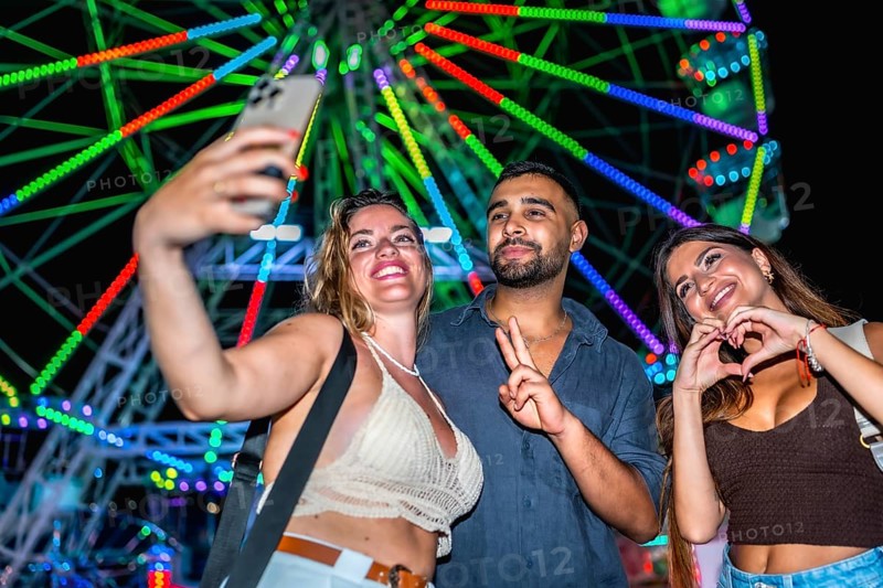 Three friends taking a selfie with smartphone in front of a colorful ferris wheel, enjoying nightlife and summer holidays