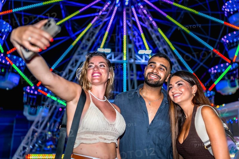 Three happy friends taking a selfie with smartphone in front of a colorful ferris wheel at an amusement park