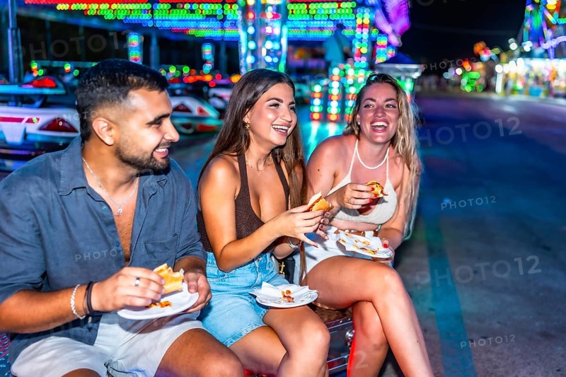 Three friends are sitting and enjoying hot dogs together at an amusement park, illuminated by colorful night lights