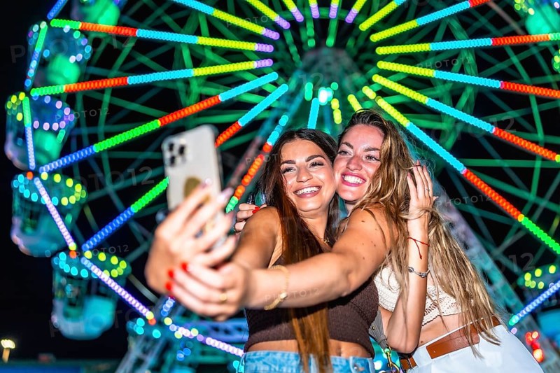 Two young women taking a selfie with a smartphone in front of a brightly lit ferris wheel at an amusement park at night, enjoying their time together