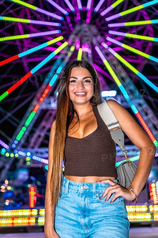 Happy young woman posing at night with illuminated ferris wheel in background, enjoying funfair attractions