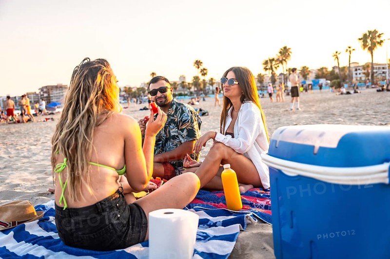 Three friends are sitting on a beach towel, enjoying a picnic with watermelon and drinks at sunset