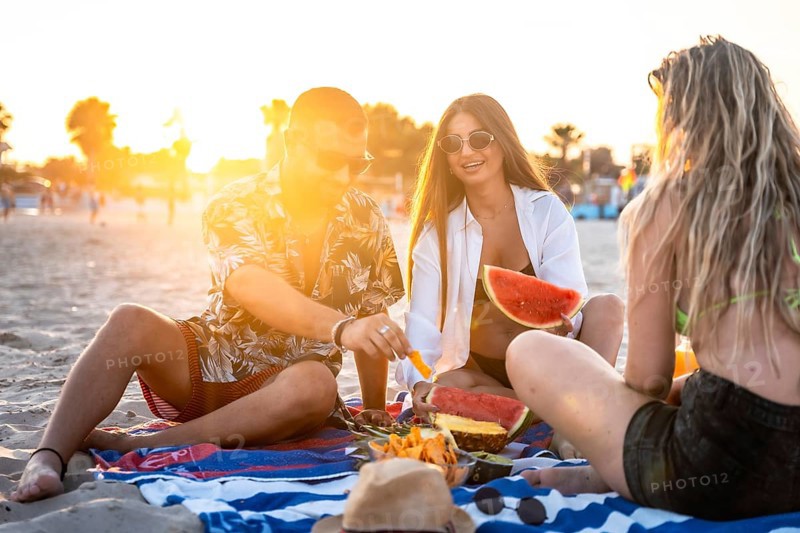 Friends sharing laughter and enjoying juicy watermelon and sweet pineapple during a picturesque sunset picnic on the beach