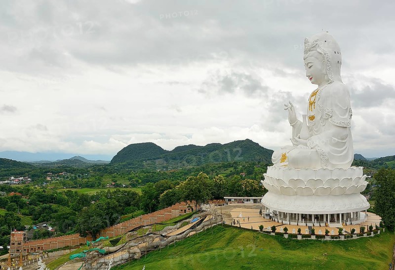 Majestic white guan yin statue sits serenely on a hilltop, overlooking a lush green valley and distant mountains in thailand