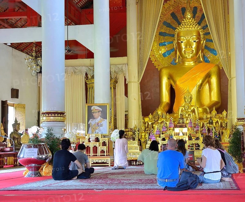 Tourists kneeling and praying in front of a giant golden buddha statue inside a buddhist temple in thailand, demonstrating reverence and cultural immersion