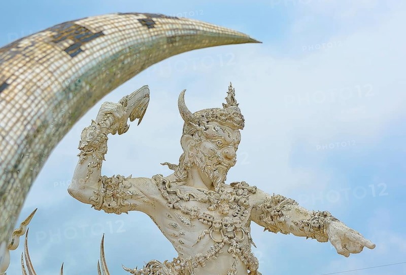 White statue of a mythological being with horns raising an arm towards a mosaic arch against a blue sky in thailand