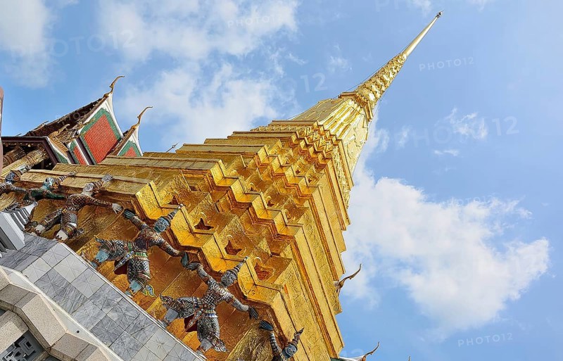 Golden chedi reaching for a bright blue sky, supported by decorative figures, within the majestic grand palace in bangkok, thailand