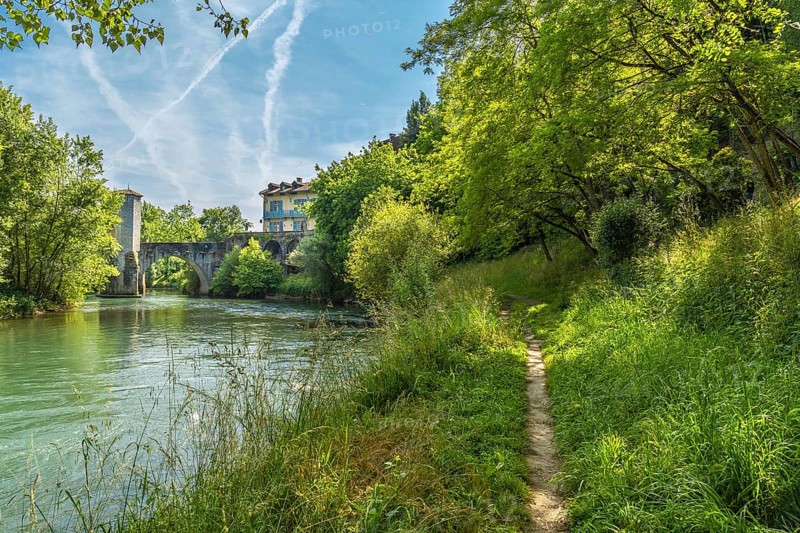 Lush vegetation frames a tranquil pathway meandering along the gave d'oloron river, leading towards the historic bridge and charming houses of sauveterre de bearn, france, under a vibrant sky