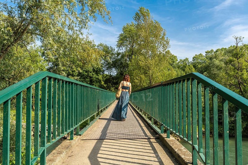 Woman walking away on a pedestrian bridge over gave d'oloron river, visiting sauveterre de bearn in france on a sunny summer day