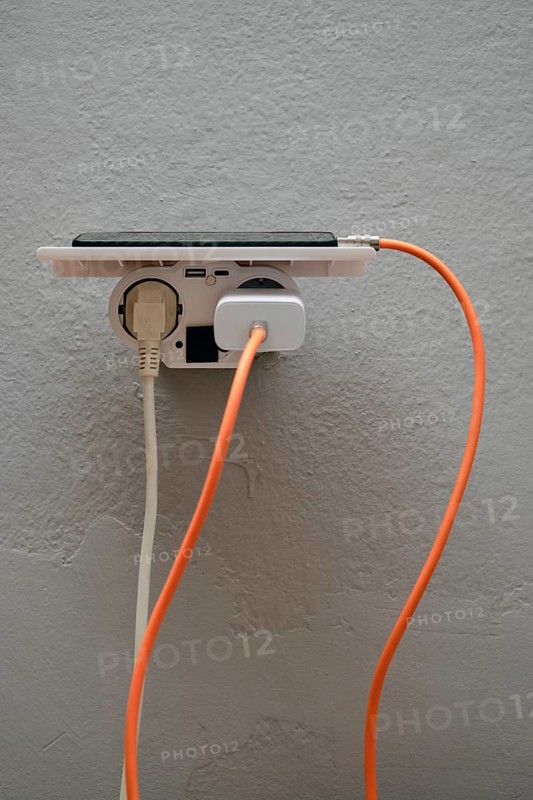 A smartphone is plugged into a wall outlet, showcasing cables in orange and white. This setup emphasizes the convenience of charging multiple devices in an indoor space, emphasizing technology use